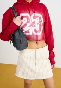 Felpa rossa corta con il numero "20" bianco e un design a stella; abbinata a una gonna beige a righe e una piccola borsa nera a tracolla.