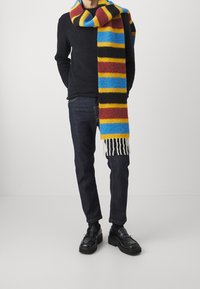 Monivärinen huivi, jossa on sinisiä, keltaisia, mustia ja punaisia vaakaraitoja, valmistettu pehmeästä, pörröisestä materiaalista, jonka päissä on hapsut.