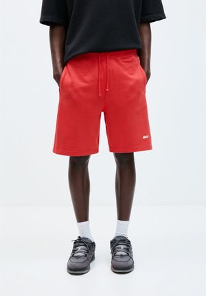 Røde bomuldsshorts med elastisk talje, sidelommer og et lille logo foran. Båret med mørke sneakers og hvide sokker.