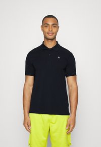 Svart bomullspolo med korte ermer, to-knapps placket og en liten hvit logo på venstre bryst. Kombinert med lyse gule shorts.