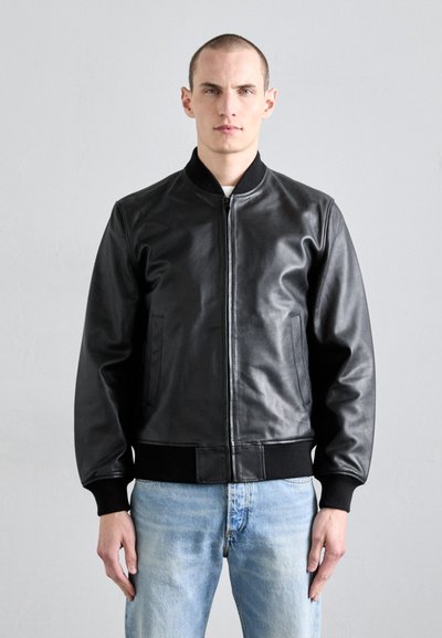 Giubbotto bomber in pelle nera con zip sul davanti, colletto e polsini a coste, tasche laterali e vita aderente. Indossato sopra jeans azzurri chiari.
