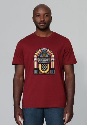 Mann trägt ein weinrotes T-Shirt mit einer bunten Vintage-Jukebox-Grafik und steht vor einem schlichten grauen Hintergrund.