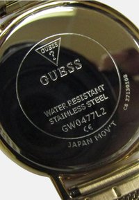 Rustfrit stål urkasse mærket "GUESS," med "VANDTÆT" og "JAPAN MOV'T." Cirkulær, blank metaloverflade med indgraveringer.