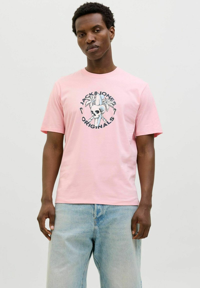 Rosa Baumwoll-T-Shirt mit einem zentralen grafischen Design, das einen Totenschädel und Palmen zeigt, rundem Ausschnitt und kurzen Ärmeln. Getragen mit hellblauen Jeans.