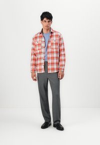 Camicia di flanella a quadri rossi e bianchi con due tasche sul petto, indossata sopra un top azzurro chiaro, abbinata a pantaloni eleganti grigi e scarpe nere.