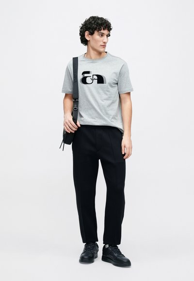 Sivé bavlnené tričko s čiernym logom, spojené s čiernymi zúženými nohavicami a čiernymi teniskami. Model drží čiernu tašku na rameno.