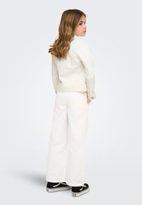 Νεαρό άτομο με κυματιστά ξανθά μαλλιά, που φοράει λευκό τζιν μπουφάν, φαρδιά λευκά παντελόνια και μαύρα αθλητικά παπούτσια, στέκεται πλάγια με τα χέρια στις τσέπες.