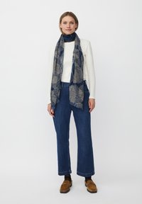 Kvinde iført en hvid langærmet top, blå mønstret tørklæde, brede blå jeans, sorte sokker og brune ruskinds loafers, stående foran en hvid baggrund.