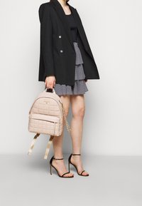 Zaino trapuntato rosa chiaro con forma ad arco, tasca frontale con zip e accentuazione a catena. Indossato con un blazer nero e una gonna stratificata.