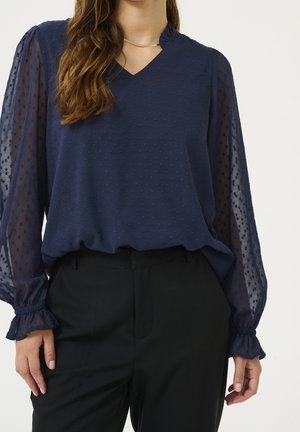 Femme portant un chemisier bleu marine texturé avec des manches transparentes à pois et un pantalon noir, debout devant un fond uni.