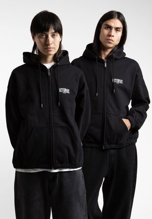 Mustad musta värvi zip-up kapuutsiga jakid, mis on valmistatud pehmest kangast, sisaldavad eesmist taskut, reguleeritavaid nööre ja logo vasakul rinnakorvil. Püksid on samuti mustad.