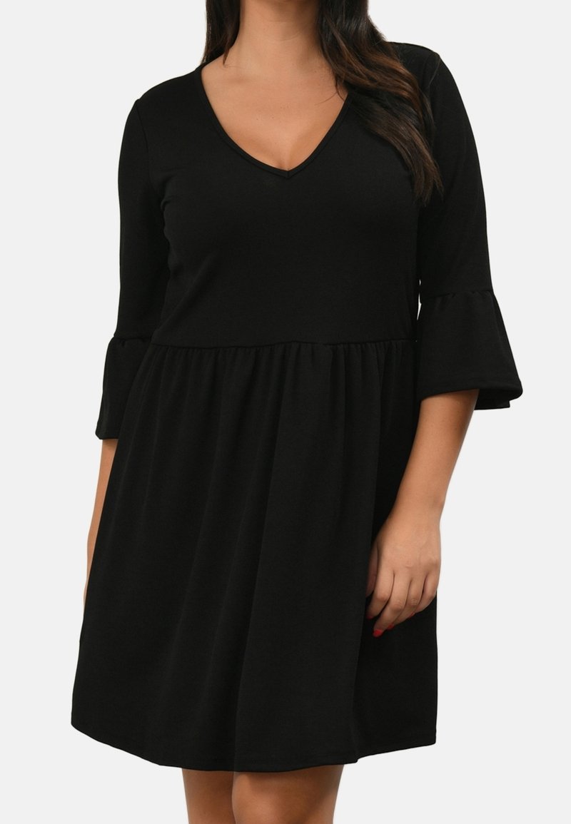 Femme portant une robe noire longueur genou avec des manches cloche trois-quarts et un décolleté en V, debout avec une main reposant à ses côtés.
