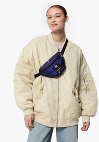 Marsupio blu con un audace motivo di testo nero, realizzato in tessuto resistente. Indossato a tracolla con una tracolla nera regolabile sopra una giacca bomber beige.