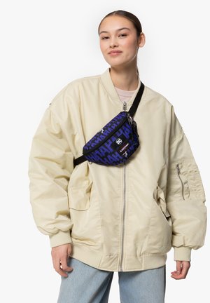 Marsupio blu con un audace motivo di testo nero, realizzato in tessuto resistente. Indossato a tracolla con una tracolla nera regolabile sopra una giacca bomber beige.