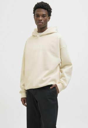 Beige hoodie with a drawstring hood, featuring embossed text on the front. Paired with black pants, smooth texture, relaxed fit.