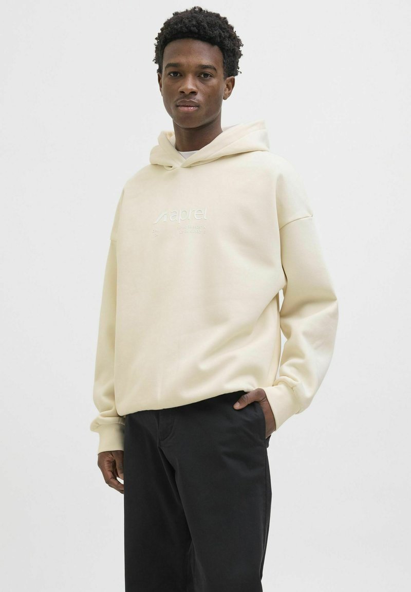 Beige hoodie with a drawstring hood, featuring embossed text on the front. Paired with black pants, smooth texture, relaxed fit.