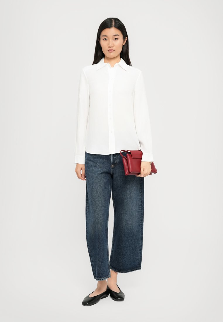Chemise blanche à manches longues et boutons, jean large bleu marine, ballerines noires, et un petit sac à main rouge tenu dans la main droite.