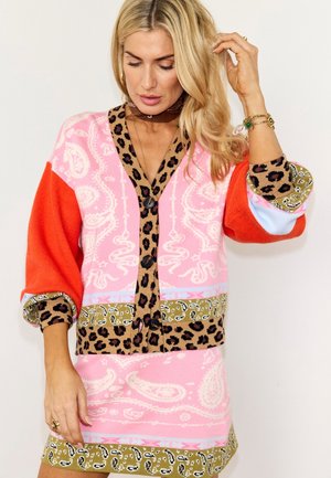 Femme portant un cardigan rose à motif cachemire et une jupe assortie avec une bordure à imprimé léopard et des manches orange vif, ajustant ses cheveux.