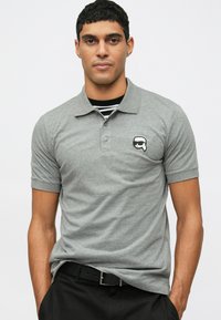 Γκρι πόλο μπλούζα με μικρό μαύρο και λευκό λογότυπο στο στήθος, κοντά μανίκια και κλασικό γιακά, συνοδευόμενη από μια μαύρη ζώνη.