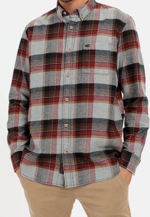 Homme portant une chemise en flanelle à manches longues boutonnée avec un motif à carreaux rouge, gris, noir et beige, et un pantalon beige, les mains dans les poches.