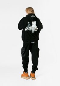 Persona con cabello rubio a la altura de los hombros que lleva una sudadera negra con una impresión de dibujos animados en la espalda, pantalones cargo negros y botas de trabajo color beige, de pie sobre un suelo blanco.