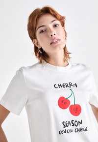 Weißes Baumwoll-T-Shirt mit einem Kirschgrafikdesign, das rote Kirschen und den verspielten Text "KIRSCHENSÄISON" zeigt. Kurze Ärmel und runder Ausschnitt.