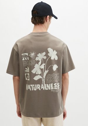 Muž nosí béžové tričko s bielymi kvetinovými a včelími grafikami a textom "NATURALNESS" na chrbte, v kombinácii s tmavou čiapkou a svetlými nohavicami.