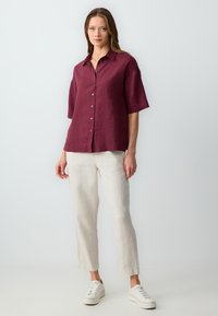Camicia di lino borgogna con maniche corte e bottoni, abbinata a pantaloni di lino beige chiaro e sneakers bianche su uno sfondo neutro.