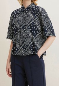 Person trägt ein marineblaues Kurzarm-Hemd mit Knopfleiste und weißem Paisleymuster sowie taillierte marineblaue Hose, eine Hand in der Tasche, Ausschnitt auf Halsniveau.