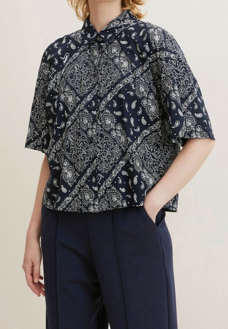 Person trägt ein marineblaues Kurzarm-Hemd mit Knopfleiste und weißem Paisleymuster sowie taillierte marineblaue Hose, eine Hand in der Tasche, Ausschnitt auf Halsniveau.