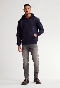 Námořnicky modrá mikina na zip s kapucí na stahovací šňůrku, předními kapsami a decentním logem, doplněná o šedé džíny a hnědé boty.