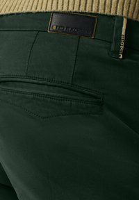 Donker groene broek heeft een gestructureerde stof met een soepele afwerking. De achterzak heeft zichtbare stiksels en een merkpatch van leer op de tailleband.