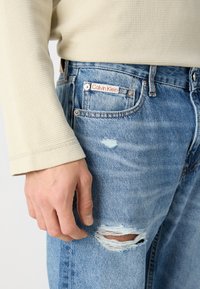 Svijetlosmeđa majica dugih rukava u kombinaciji sa svijetloplavim trapericama koje imaju potrgani izgled i vidljiv Calvin Klein logo na džepu.