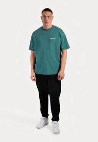 Blågrøn bomulds T-shirt med rund hals og korte ærmer, med et diskret hvidt logo. Kombineret med sorte bukser og hvide sneakers.