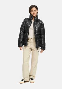 Black puffer jacket with a high collar, glossy finish, front zipper, and large pockets, paired with light-colored pants and sneakers.