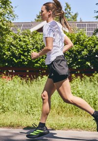 Schwarze Laufshorts kombiniert mit einem weißen Sport-T-Shirt und schwarzen Sneakers mit grünen Streifen, die ein aerodynamisches Design aufweisen.