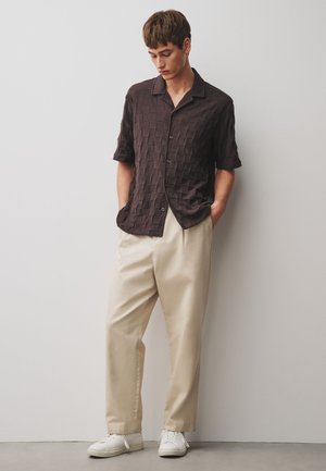 Young man wearing a dark brown textured short-sleeve button-up shirt, beige trousers, and white sneakers, standing against a plain wall.