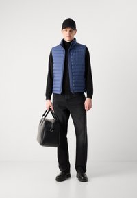 Chaleco azul acolchado, suéter de cuello alto negro, jeans oscuros y botas negras; sosteniendo un bolso de cuero negro con dos asas. Fondo liso.