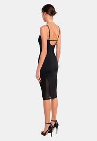 Schwarzes, figurbetontes Kleid mit verstellbaren Spaghettiträgern, durchsichtigen Seitenteilen, knielangem Saum und enger Silhouette. Träger mit schwarzen High Heels.