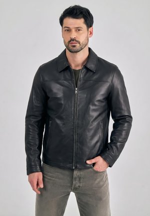 Hombre con cabello corto y oscuro y barba, que lleva una chaqueta negra de cuero con cremallera y jeans grises, de pie frente a un fondo liso.