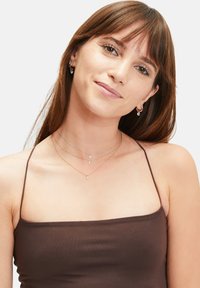 Schichtketten mit metallischen Ketten in Silber und Ros égold, die tropfenförmige Anhänger tragen. Einfache Creolen ergänzen den Look.