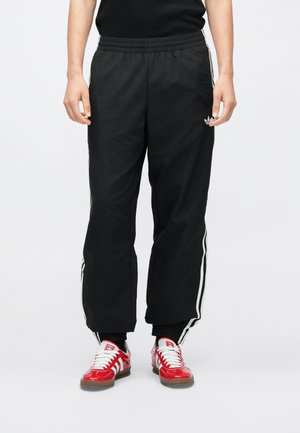 Person trägt schwarze Adidas-Trainingshose mit weißen Seitenstreifen und roten Adidas-Sneakern und steht vor einem weißen Hintergrund.
