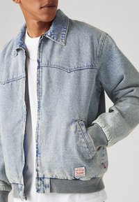 Veste en denim bleu clair avec deux poches poitrine, des poignets et un ourlet côtelés, fermeture éclair et une petite étiquette sur le côté inférieur gauche.