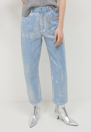 Person in hellblauen, metallisch schimmernden Jeans und silbernen, spitz zulaufenden Knöchelschuhen vor einem schlichten weißen Hintergrund.