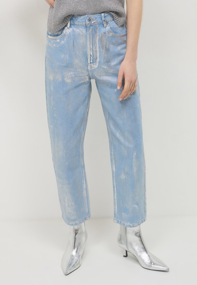 Persona che indossa jeans blu chiaro con finitura metallica e stivaletti argento a punta su uno sfondo bianco uniforme.