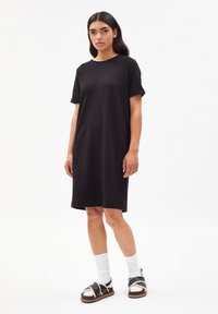 Robe T-shirt en coton noir avec une coupe décontractée, manches courtes et longueur au genou. Portée avec des sandales noires et des chaussettes blanches. Texture unie.