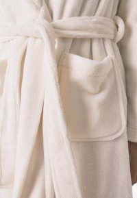 Biały pluszowy szlafrok o miękkiej teksturze, z wiązanym paskiem i naszywaną kieszenią z przodu. Materiał wydaje się gruby i przytulny.