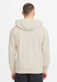 Felpa beige con cappuccio regolabile, texture morbida e polsini a coste. Presenta un piccolo etichetta visibile nella cucitura laterale. Indossata con pantaloni scuri.