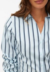 Jasnoniebieska koszula na guziki z pionowymi granatowymi pasami i cienkimi białymi akcentami. Materiał wydaje się gładki, z klasycznym kołnierzykiem.