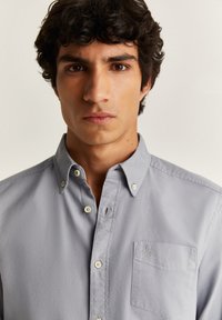 Camicia button-up grigia chiara in cotone, con colletto, taschino sul petto e un ricamo del logo discreto. Tessuto liscio e vestibilità regolare.